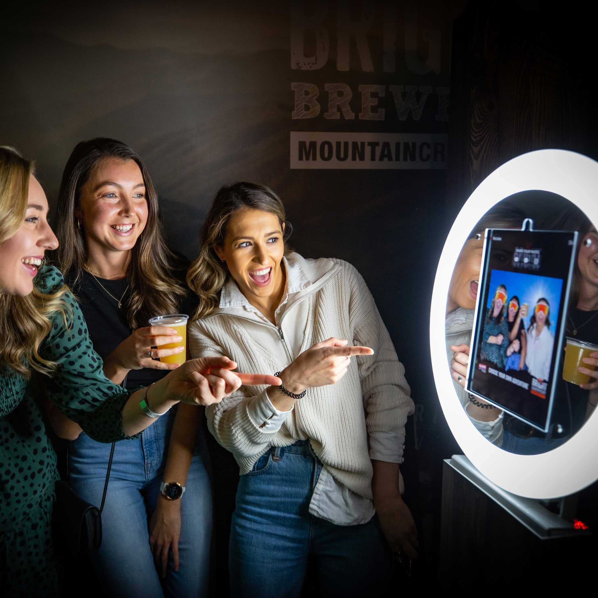 three women pointing at the photo they have just taken on the photo booth of themselves with green screen and ski masks on as a digital prop